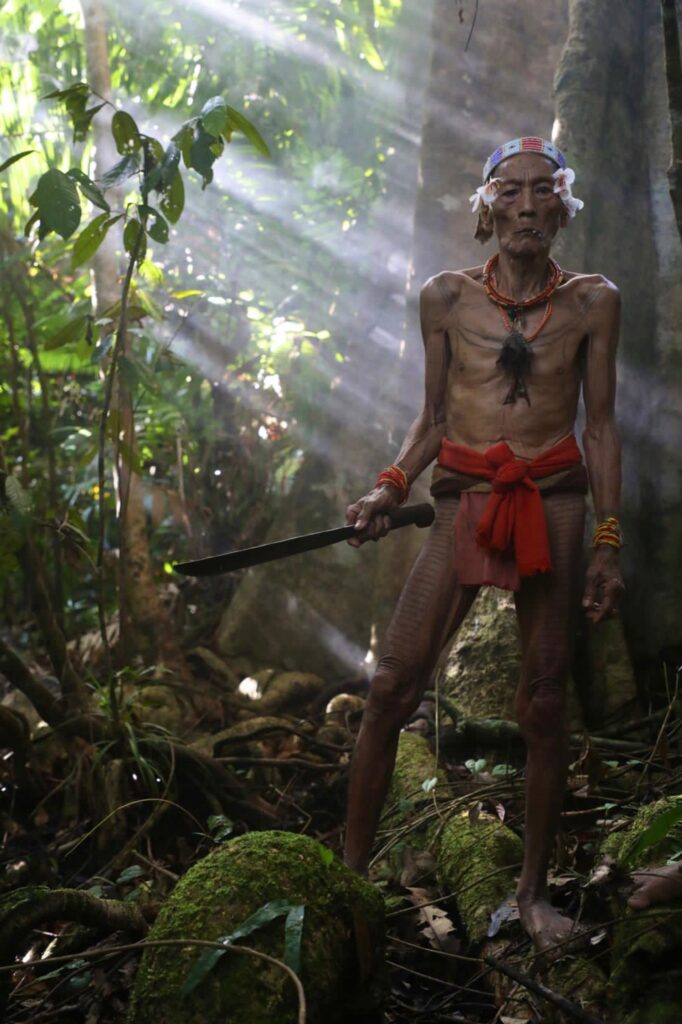 An elderly man stands in a sunlit West Sumatra rainforest, holding a machete.