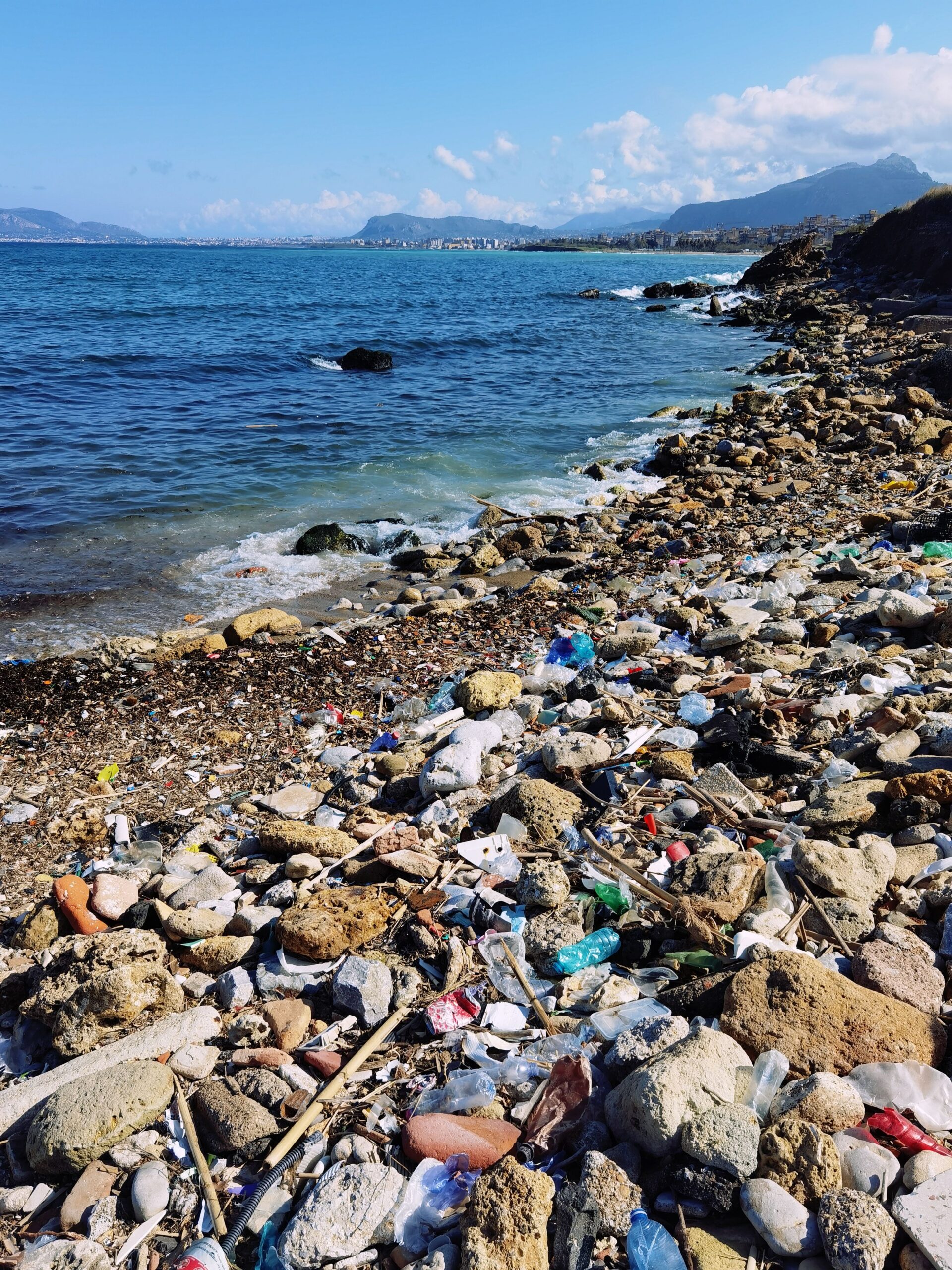 Plastic waste and debris litter a rocky beach in Sicily, highlighting environmental pollution.