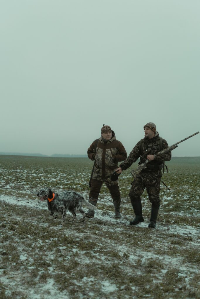 Two hunters in camouflage with a dog in a snowy field during winter.