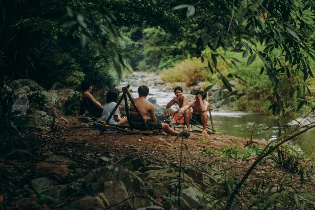 A group of adults relaxing and eating by a riverbank surrounded by lush greenery.